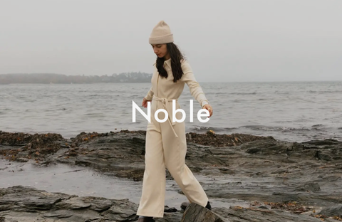 Woman standing on a rocky beach, wearing loose, comfortable clothing by Noble, symbolizing simplicity and freedom.